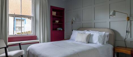 A bedroom with a bed, a chair, a window with curtains, a red bookshelf, and a wall-mounted lamp.