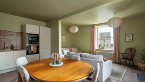 A modern kitchen with built-in appliances, a dining area with a wooden table and chairs, and a living room with a sofa and a large window. - Ledger's Cottage- Harbourfront Home Pittenweem (Pittenweem)