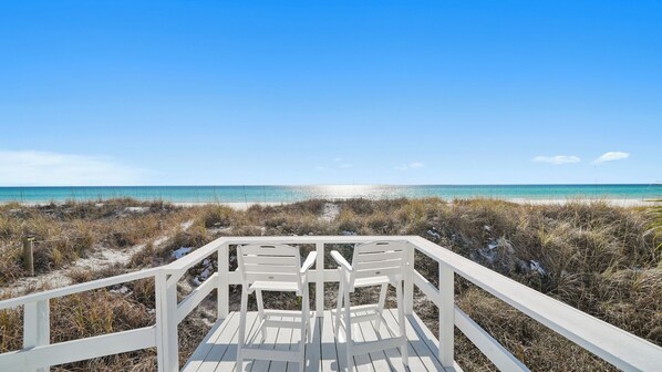 A wooden deck with white railings overlooking a beach with clear blue water.