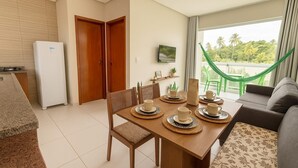 Uma área de refeições com uma mesa de madeira posta para quatro pessoas, um sofá e uma rede perto de uma grande janela com vista para palmeiras.