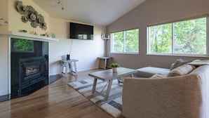 A living room with a fireplace, a sofa, a coffee table, and large windows offering a view of trees. - The Last Frontier Lake House Lodge in Anchorage 3bdrm 2ba (Anchorage)