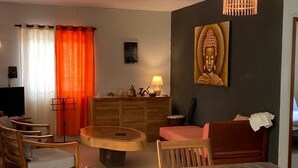 Una sala de estar con una mesa redonda de madera, un sofá y una mesa de comedor con sillas. Se observa un cuadro de Buda en la pared y una lámpara sobre una mesita auxiliar.
