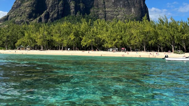 Una playa con aguas turquesas cristalinas, una gran isla rocosa al fondo y densos árboles verdes bordeando la orilla.