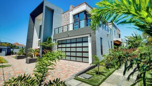 A modern two-story house with a large garage, a stone facade, and a well-maintained garden with various plants. - Luxury Oceanfront Villa~ Fully Staffed~Pool~Jacuzzi~Movie Theatre (tower isle)