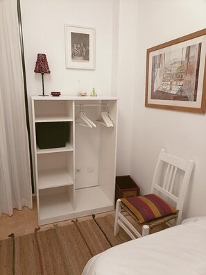 Ein Schlafzimmer mit Bett, Sessel, Kleiderschrank mit Kleiderbügeln, einem kleinen Tisch und einem Bild an der Wand.