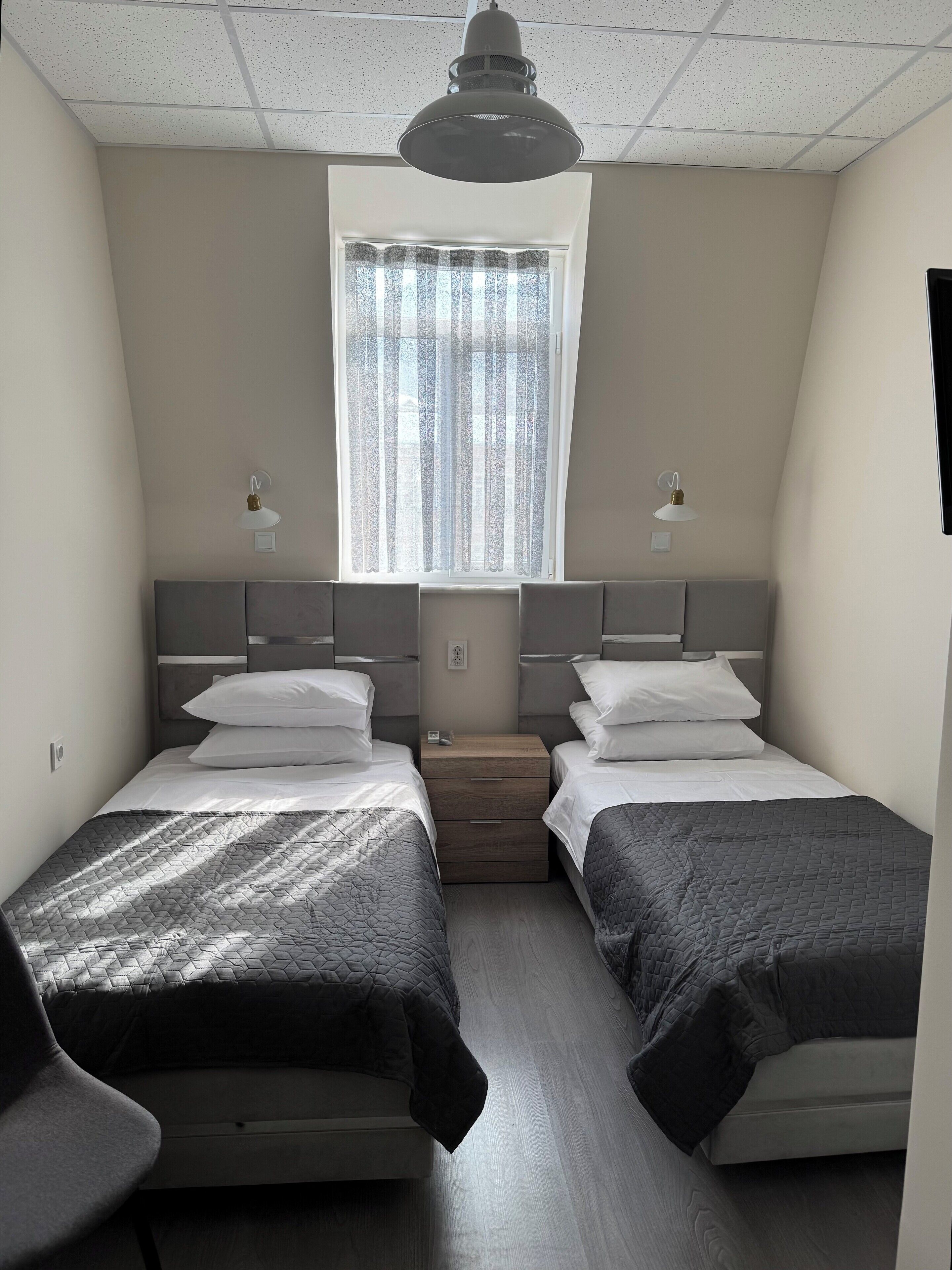 A small, modern room with two single beds, a small table, and a window with sheer curtains.