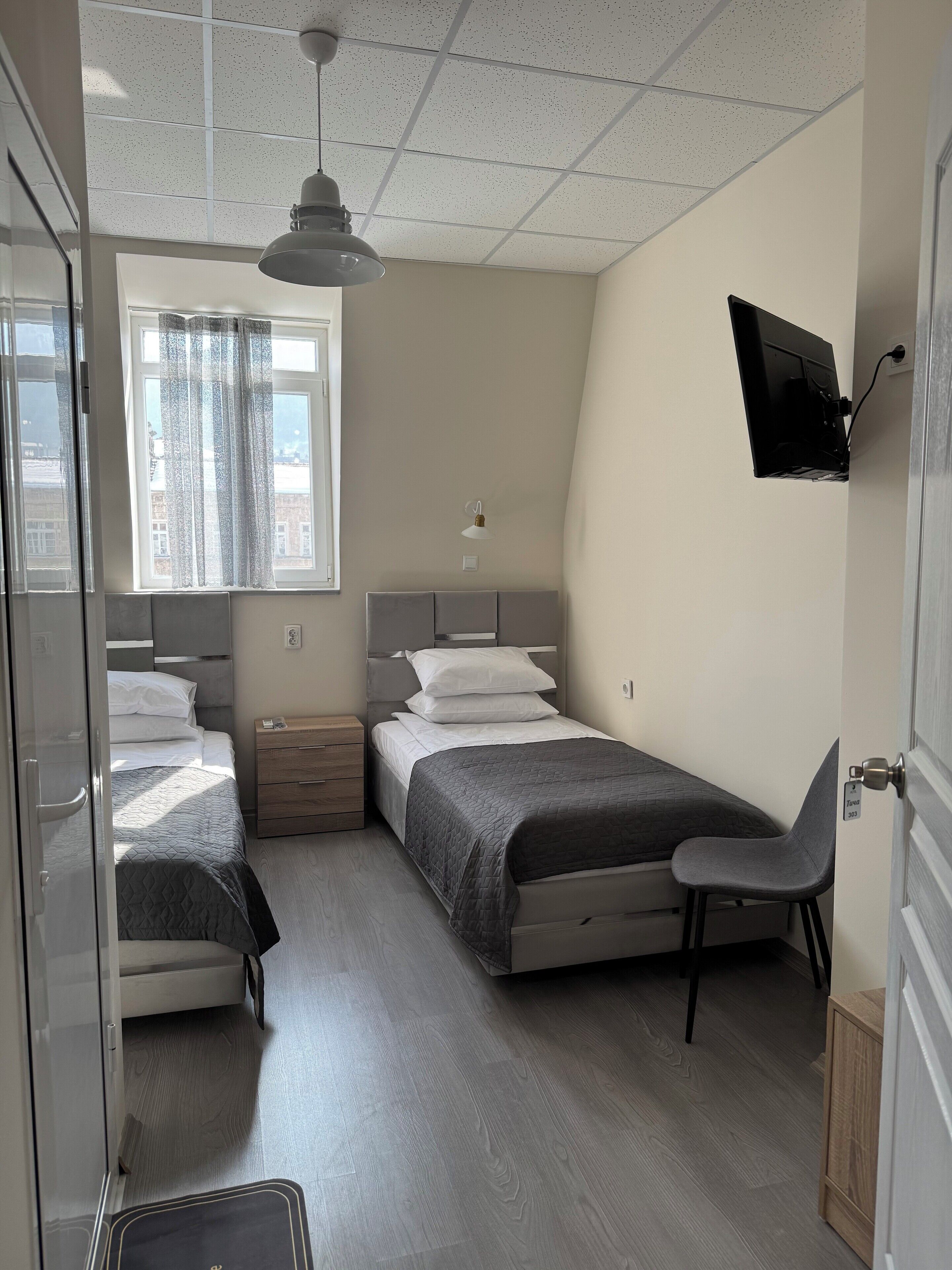 A bedroom with two beds, a desk, a chair, a TV mounted on the wall, and a window with curtains.