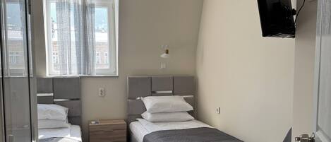 A bedroom with two beds, a desk, a chair, a TV mounted on the wall, and a window with curtains.