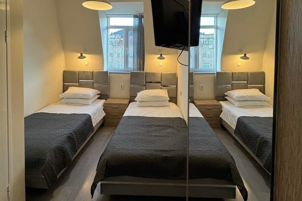 A hotel room with three single beds, each with a headboard and a bedside table. There are two pendant lights hanging from the ceiling, and a flat-screen TV mounted on the wall.