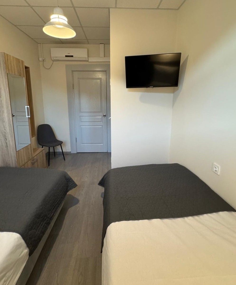 A room with two beds, a chair, a TV mounted on the wall, and an air conditioning unit.