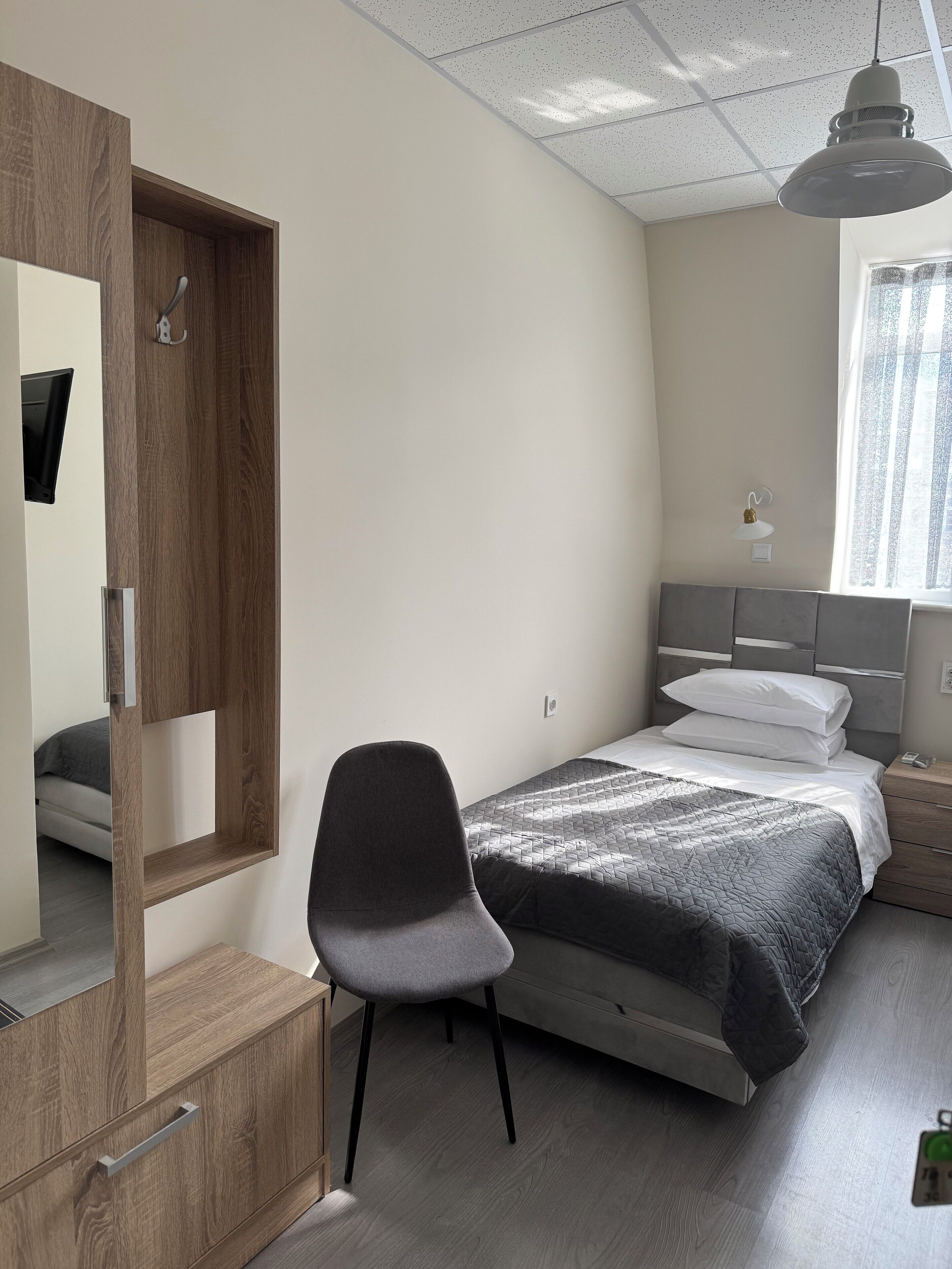 Un dormitorio con una cama, una silla, un armario de madera, un espejo, una mesita de noche y una ventana.