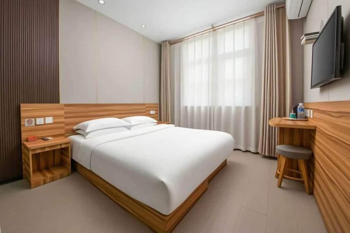 Hotelkamer met een groot bed, houten hoofdeinde, nachtkastje, bureau met stoel, tv aan de muur en raam met gordijnen.