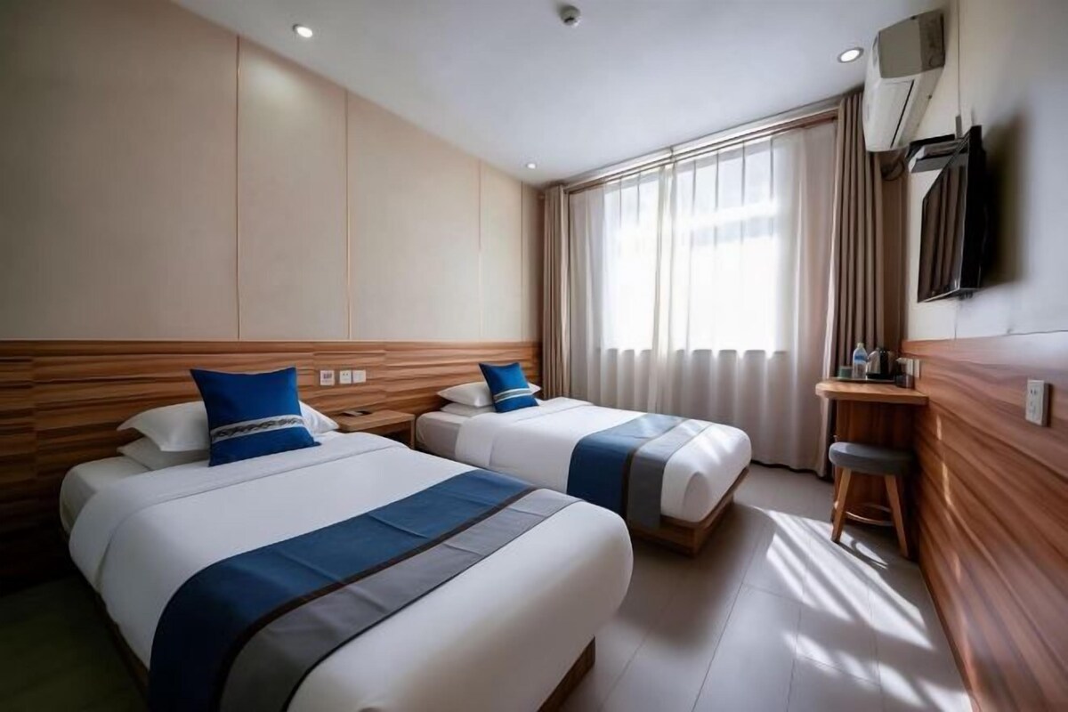 Une chambre d’hôtel avec deux lits, une télévision, un bureau avec une chaise et une grande fenêtre avec des rideaux.