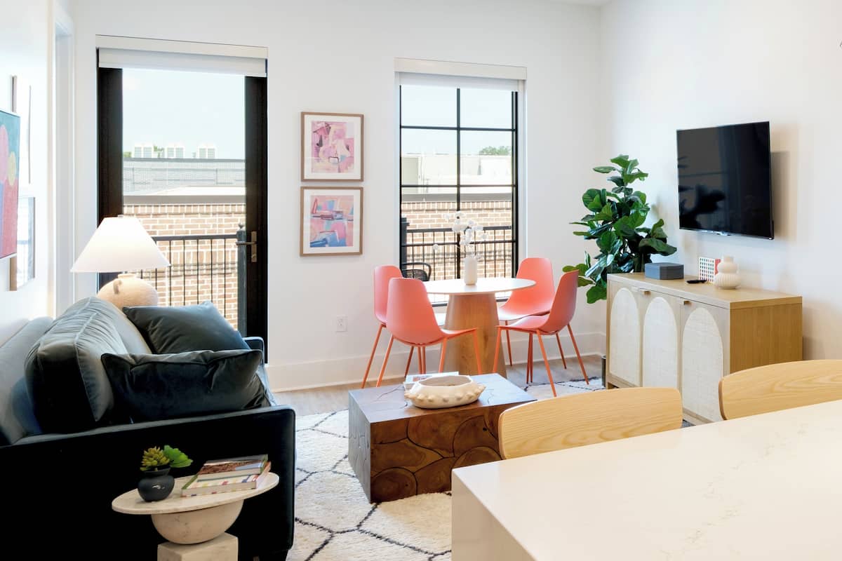 A modern living room with a sofa, a dining table with chairs, a TV mounted on the wall, and a balcony with a view.