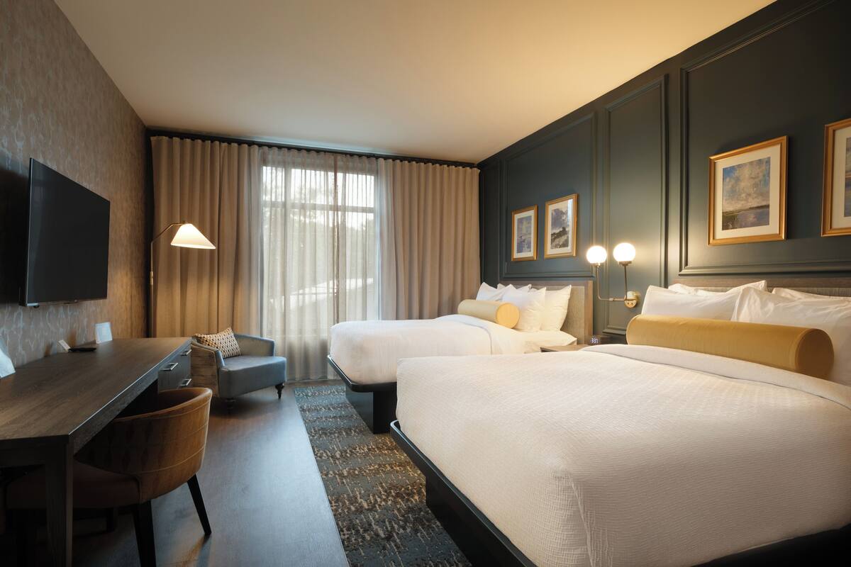 A hotel room with a large bed, a smaller bed, a desk with a chair, a TV, and a window with curtains.