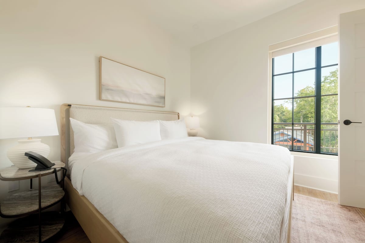 A neatly made bed in a room with a window, a bedside table with a lamp, and a framed picture on the wall.