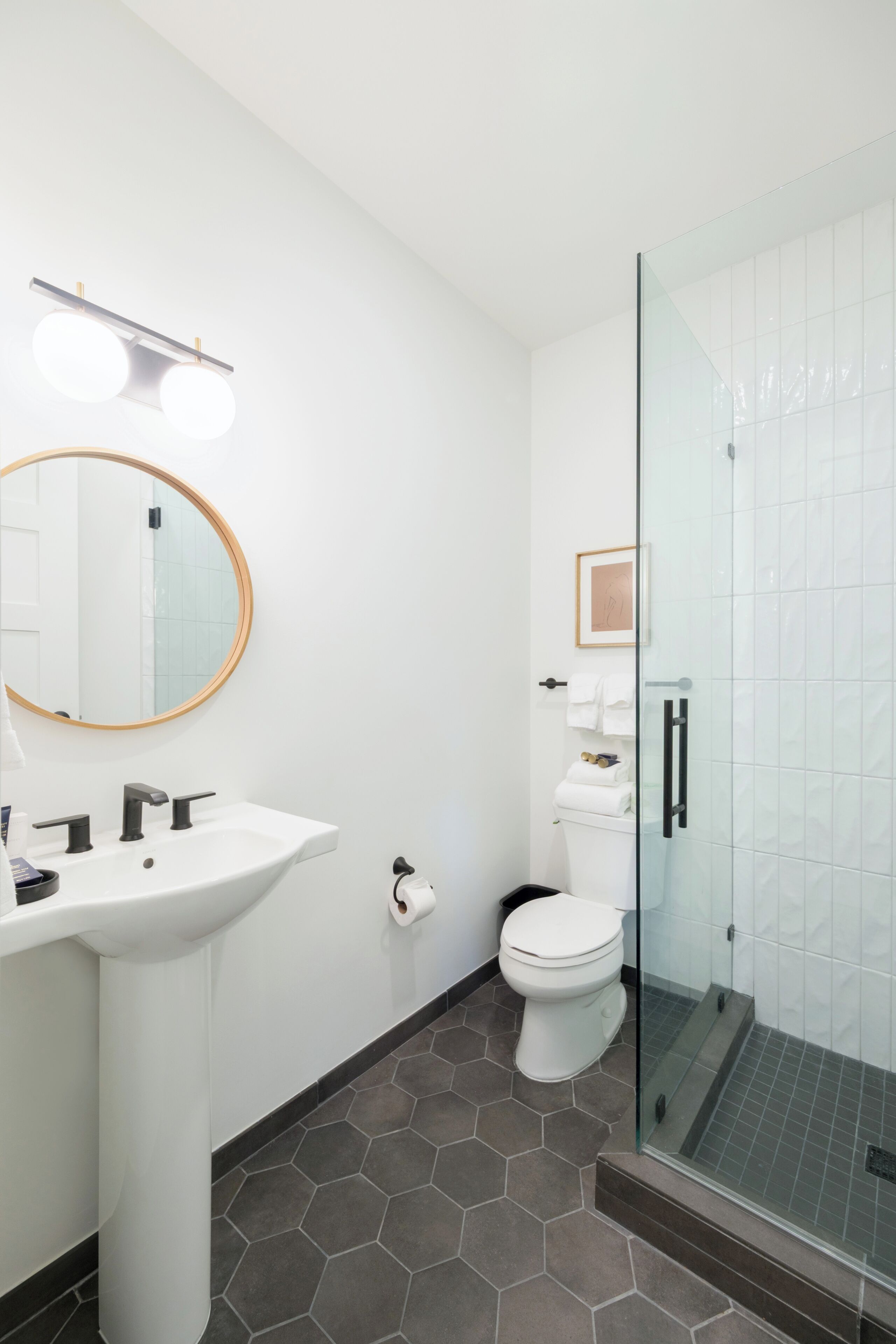 A bathroom with a white sink, round mirror, hexagonal floor tiles, a toilet, and a glass-enclosed shower.