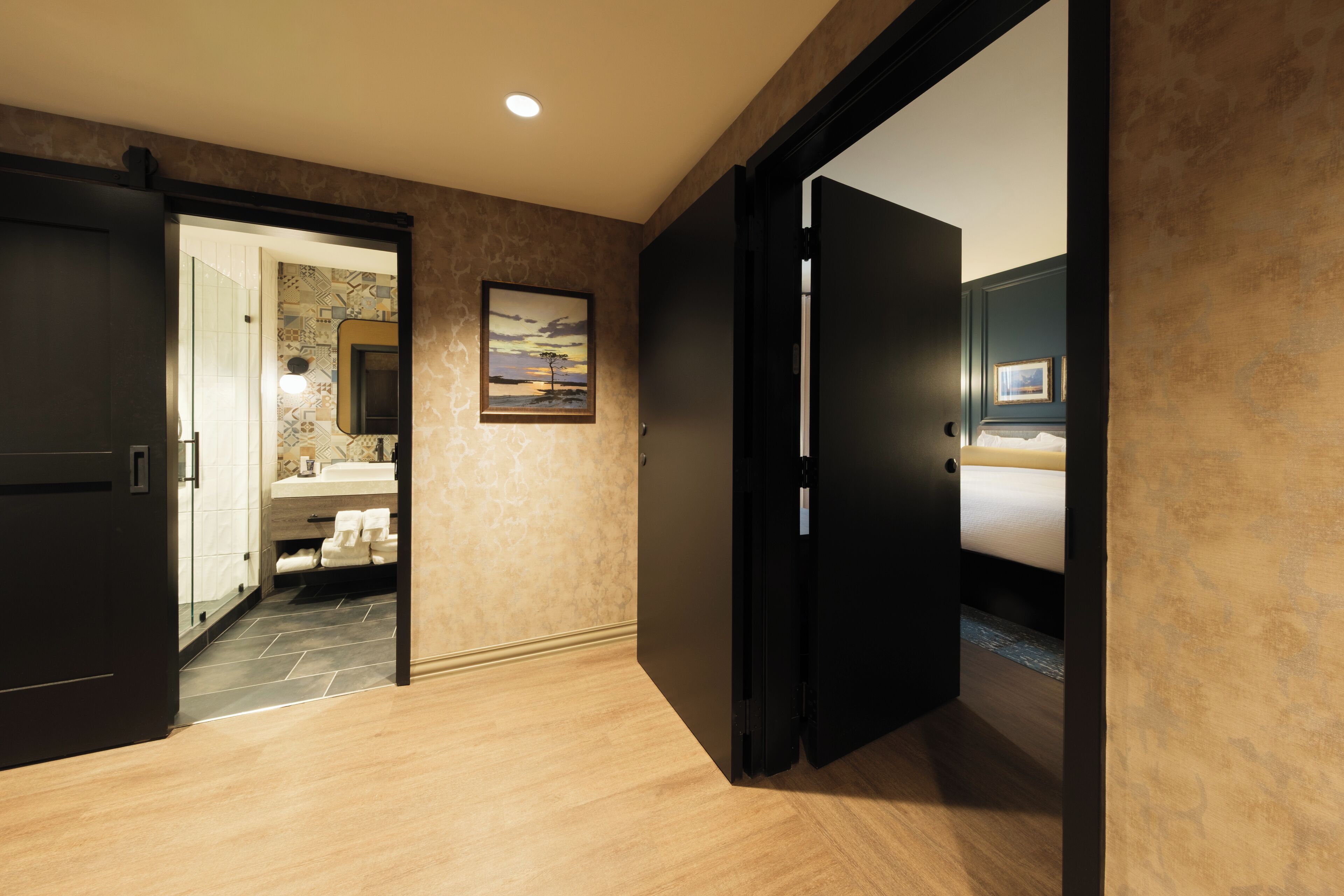 Habitación de hotel con piso de madera, baño con lavamanos y perchero, y cama con mesita de noche.