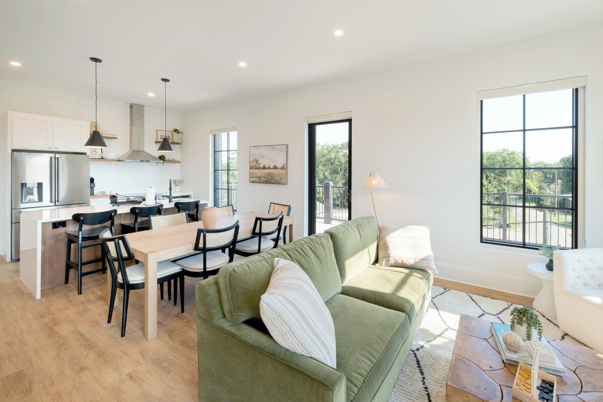A modern kitchen with a dining area, a green sofa, and a large window with a view.