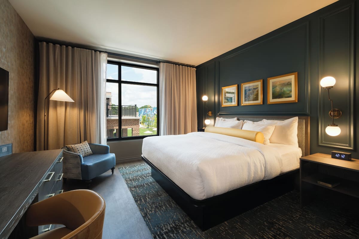 A hotel room with a large bed, a desk, a chair, a nightstand, and a view of the outdoors.