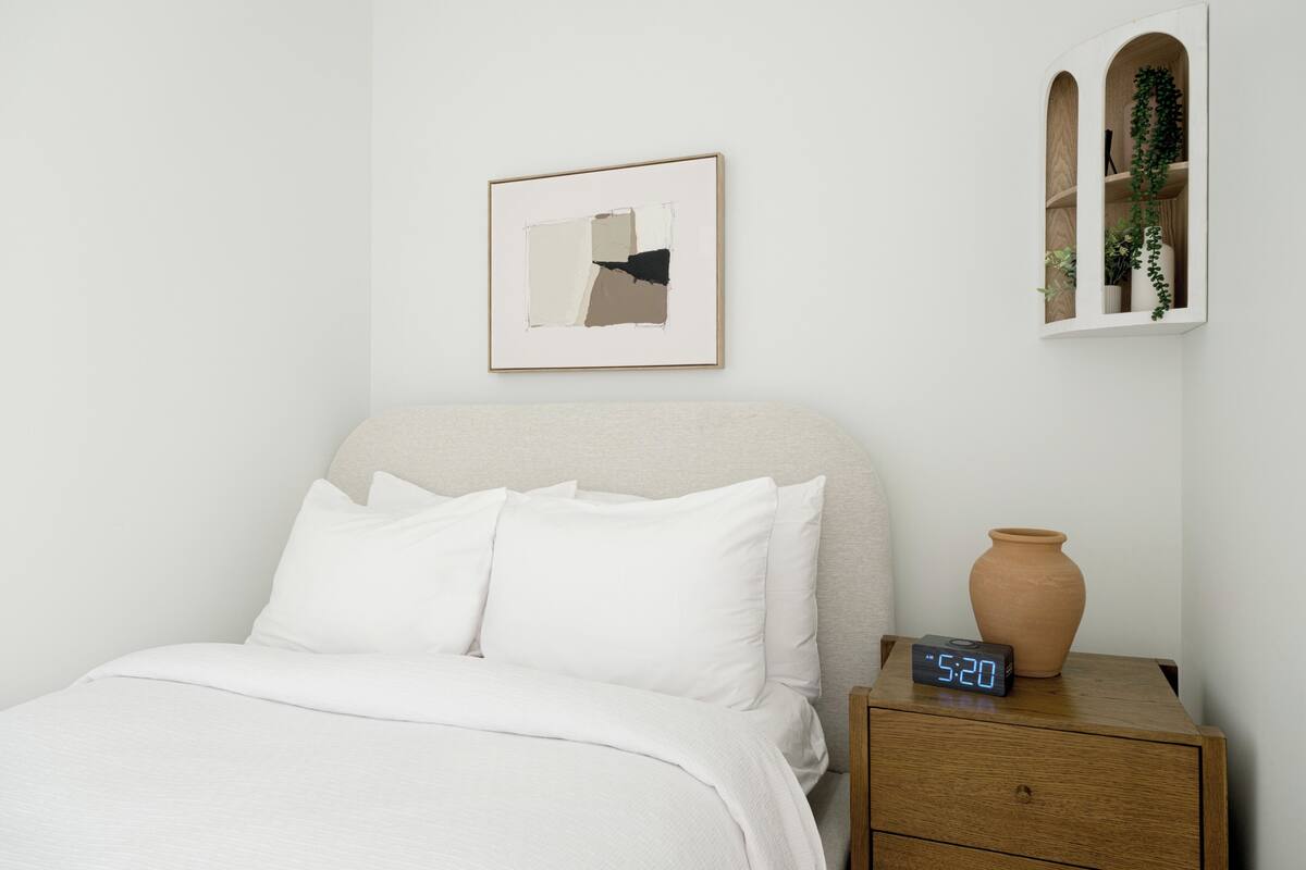A neatly made bed with white bedding, a digital clock, a framed abstract artwork, and a window with a plant.
