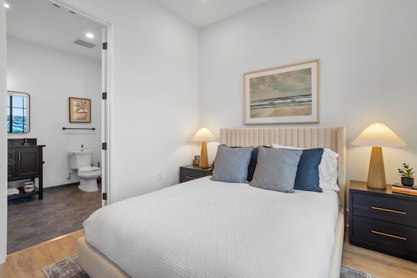 Un dormitorio con cama, mesitas de noche, un tocador y un baño visible a través de una puerta abierta.