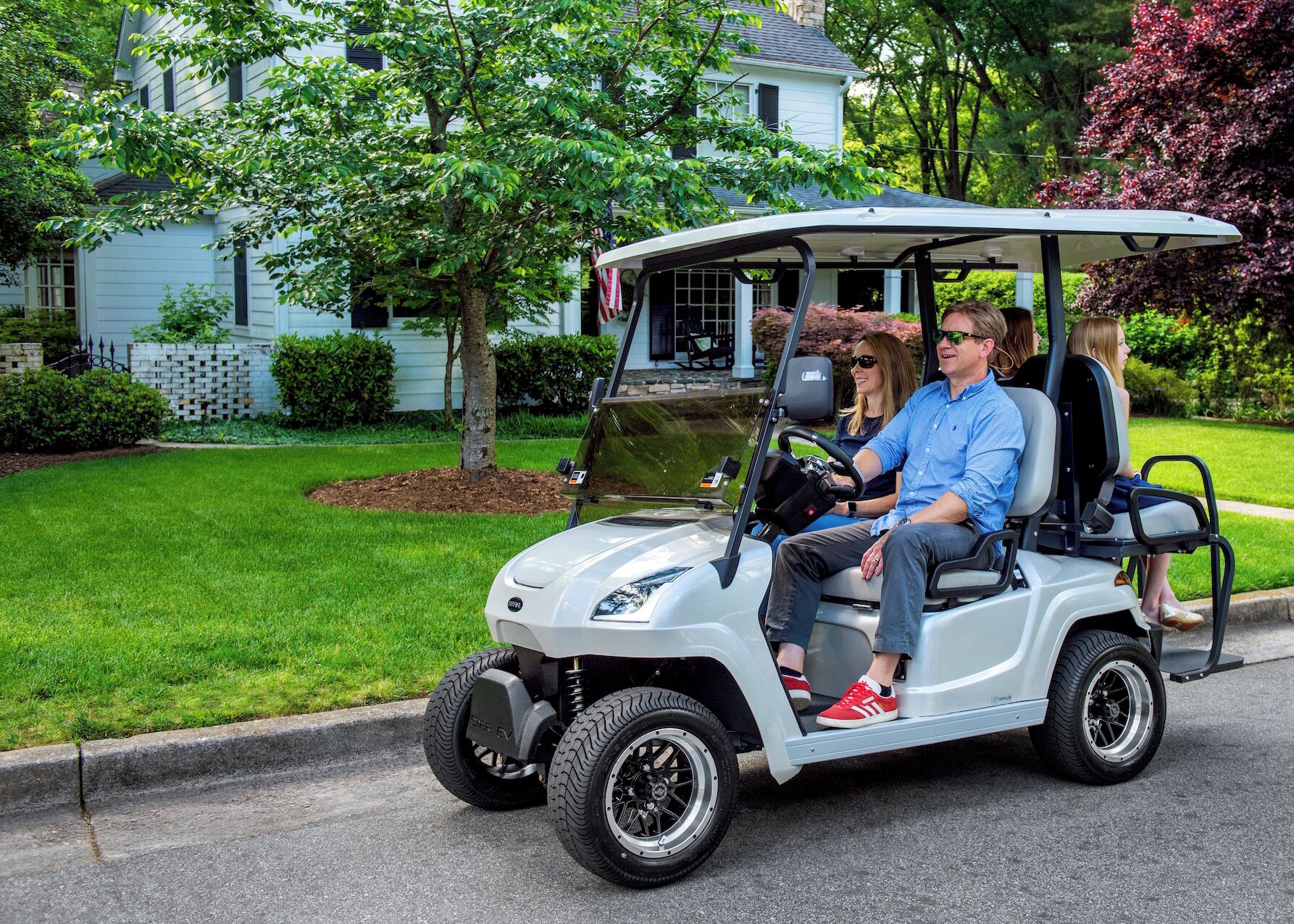 A golf cart on a residential street with a house and trees in the background.