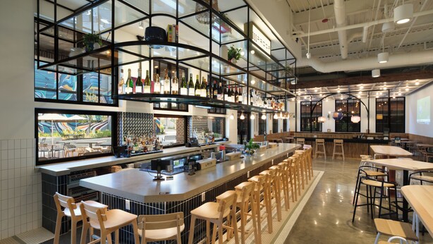 A bar area with a long counter, multiple stools, and a high ceiling with exposed beams. - OS Hotel  (Ocean Springs)