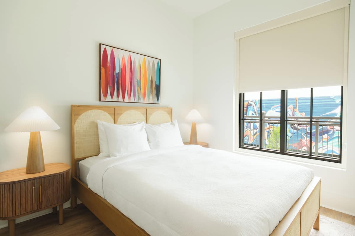 A hotel room with a bed, two bedside tables, a window with a view, and a colorful abstract painting on the wall.