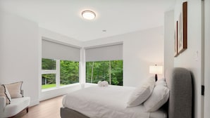 Une chambre moderne avec un grand lit, un fauteuil et une vue sur la verdure grâce à de grandes fenêtres.