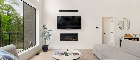 A modern living room with a large window, a fireplace, a flat-screen TV, and a round coffee table with magazines.