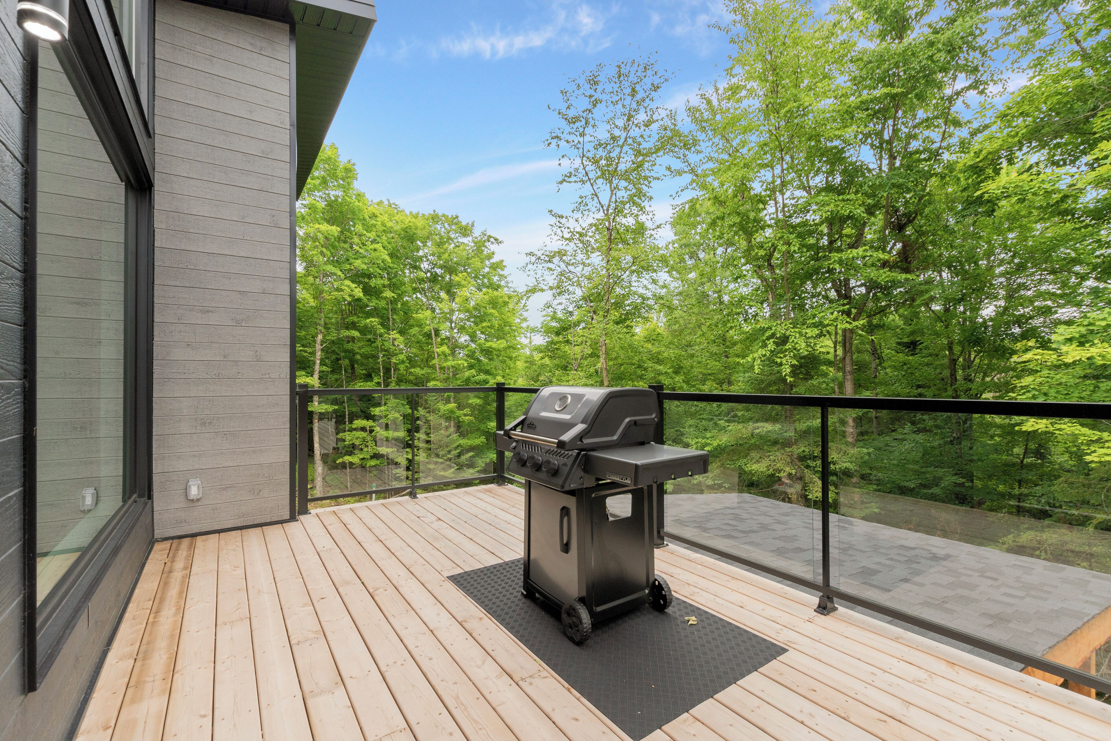 A modern deck with a built-in grill, glass railings, and a view of a forested area.