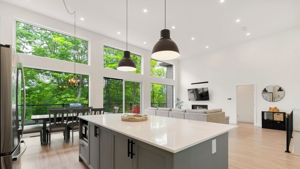 Une cuisine moderne dotée d’un grand îlot central, d’un coin repas et offrant une vue sur une végétation luxuriante grâce à de grandes fenêtres.