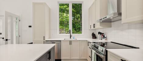 A modern kitchen with white cabinetry, a large window with a view of greenery, a built-in oven, and a stainless steel range hood.
