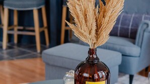 Une table ronde sur laquelle se trouvent un vase en verre foncé contenant des herbes des pampas séchées, un petit bocal en verre et un magazine.