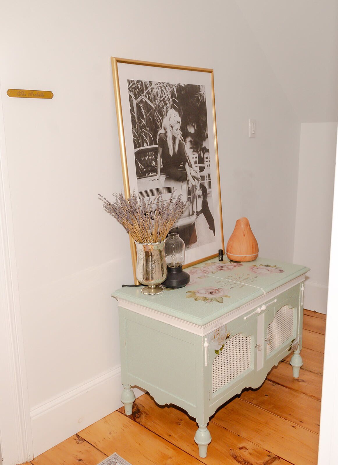 Une table de style vintage ornée d’un motif floral, une photographie en noir et blanc encadrée, un vase contenant des fleurs séchées et un petit vase orange.