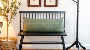 Une pièce avec un plancher en bois, un banc en métal noir agrémenté d’un coussin vert, un porte-manteau fixé au mur, un tableau encadré représentant un taureau et un panier en osier.