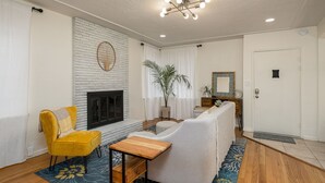Un salon avec une cheminée, un fauteuil jaune, un canapé, une petite table en bois et un tapis à motifs.