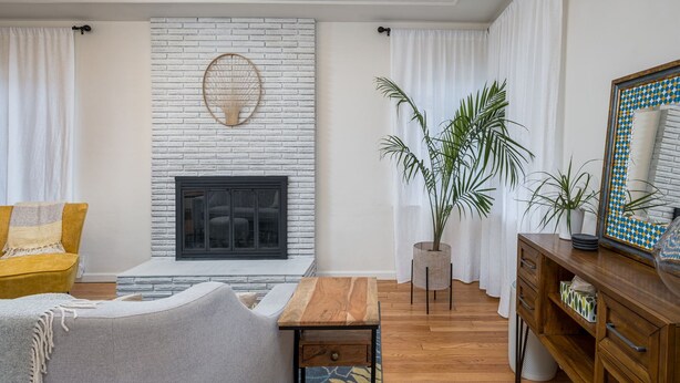 Un salon avec une cheminée en briques blanches, un canapé jaune, un fauteuil gris, une table basse en bois et une plante en pot.