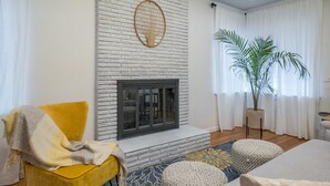Un salon avec une cheminée, un fauteuil jaune, une grande plante et un tapis à motifs.