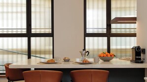 Una cocina moderna con una isla central, dos sillas marrones, una cafetera, un bol de naranjas y un ventanal con persianas.