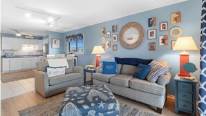 Una sala de estar con un sofá gris, un otomán estampado y una pared azul decorada con fotos enmarcadas y un espejo.