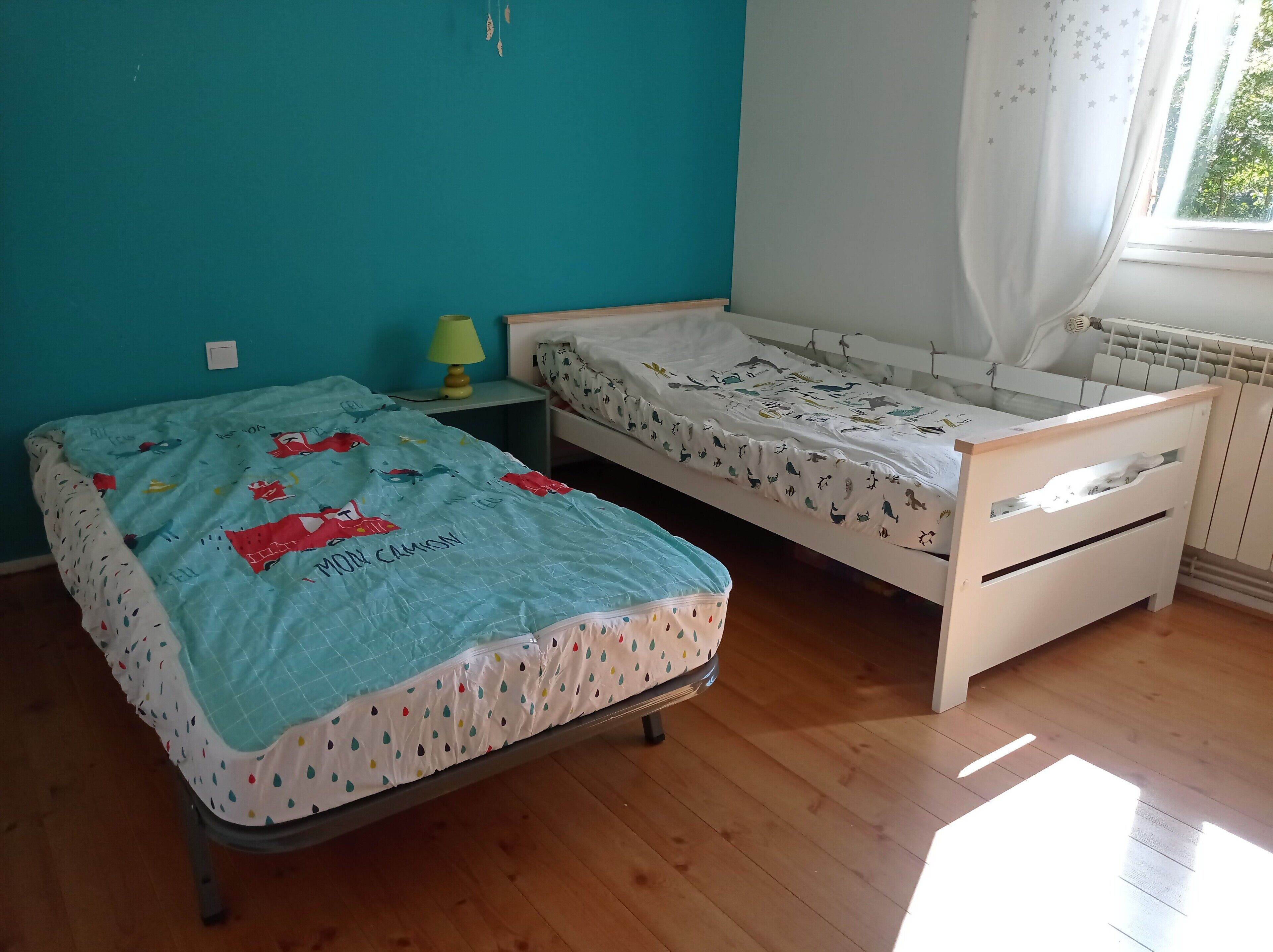 A bedroom with two single beds, a nightstand with a lamp, a window with curtains, and a radiator.