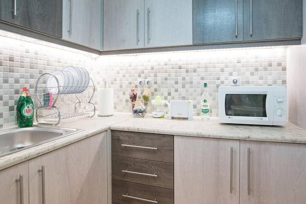 Una cocina con gabinetes grises, salpicadero de azulejos blancos, un escurreplatos, un microondas y una botella de detergente Fairy.