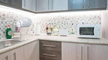A kitchen with grey cabinets, a white tiled backsplash, a dish rack, a microwave, and a bottle of Fairy dish soap.