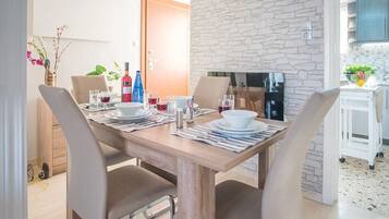 A dining area with a wooden table set for four, surrounded by chairs. A stone wall and a kitchen area are visible in the background.