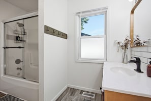 A bathroom with a glass shower enclosure, a white sink with a black faucet, a window with a view, and a wooden towel rack. - Spacious Group Home with Arcade (Champaign)