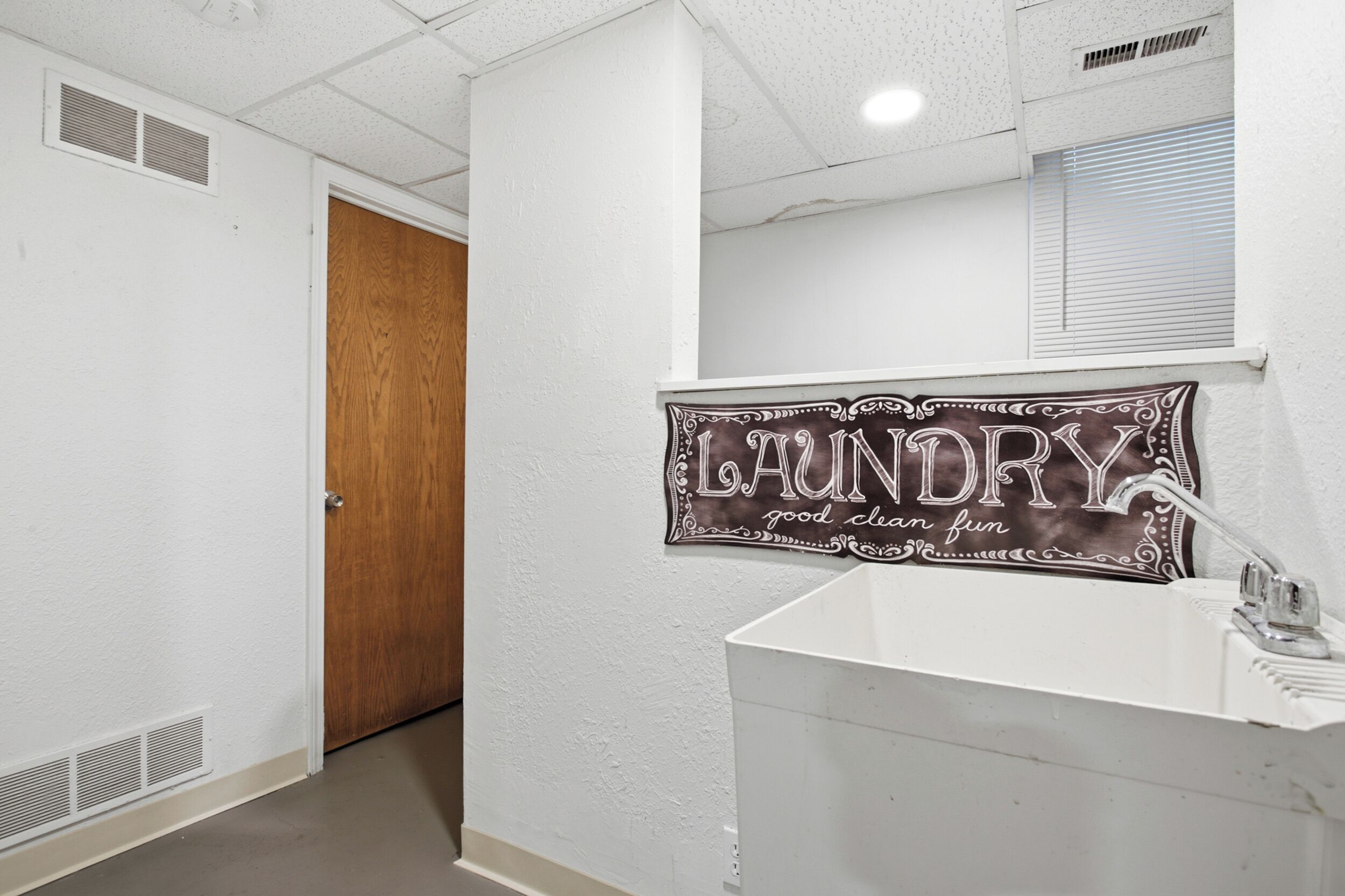 A bathroom with a white sink, a wooden door, and a wall-mounted sign that reads 'LAUNDRY good clean fun'.