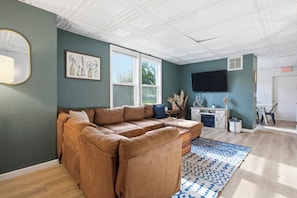 A living room with a sectional sofa, a flat-screen TV, a window, and a small table with decorative items. - Spacious Group Home with Arcade (Champaign)