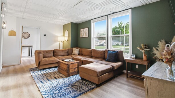 A living room with a sectional sofa, a wooden coffee table, a patterned rug, and a view of a house through large windows. - Spacious Group Home with Arcade (Champaign)